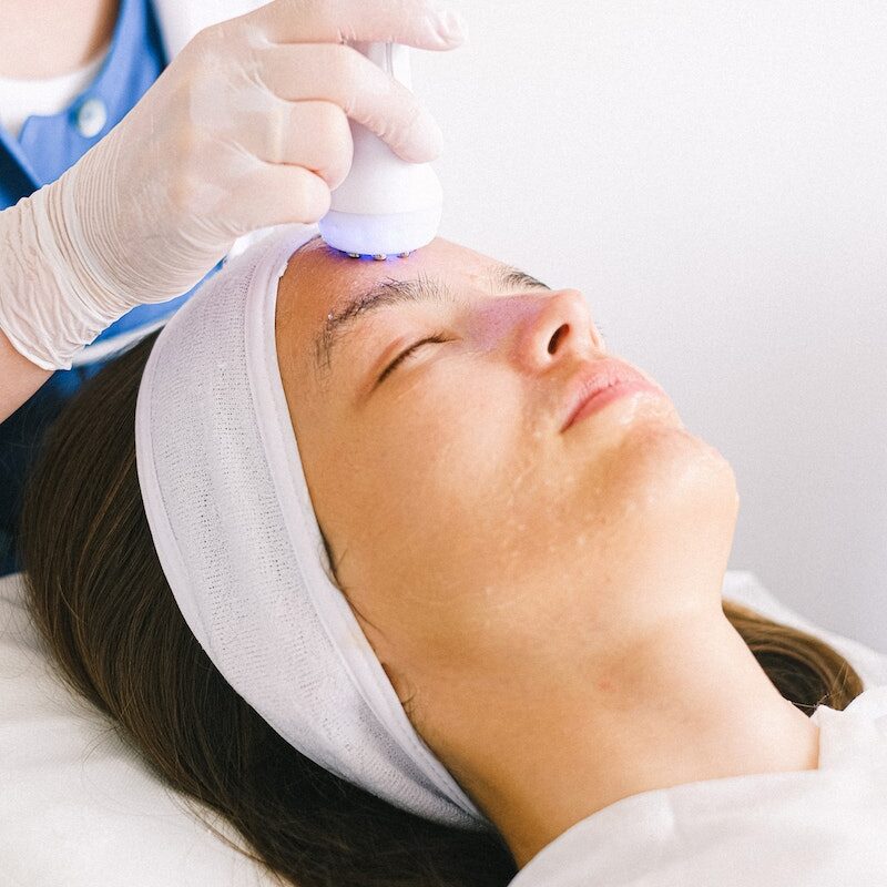 Professional female beautician in gloves and robe using ultrasound apparatus to clean female customer face lying with eyes closed in spa center